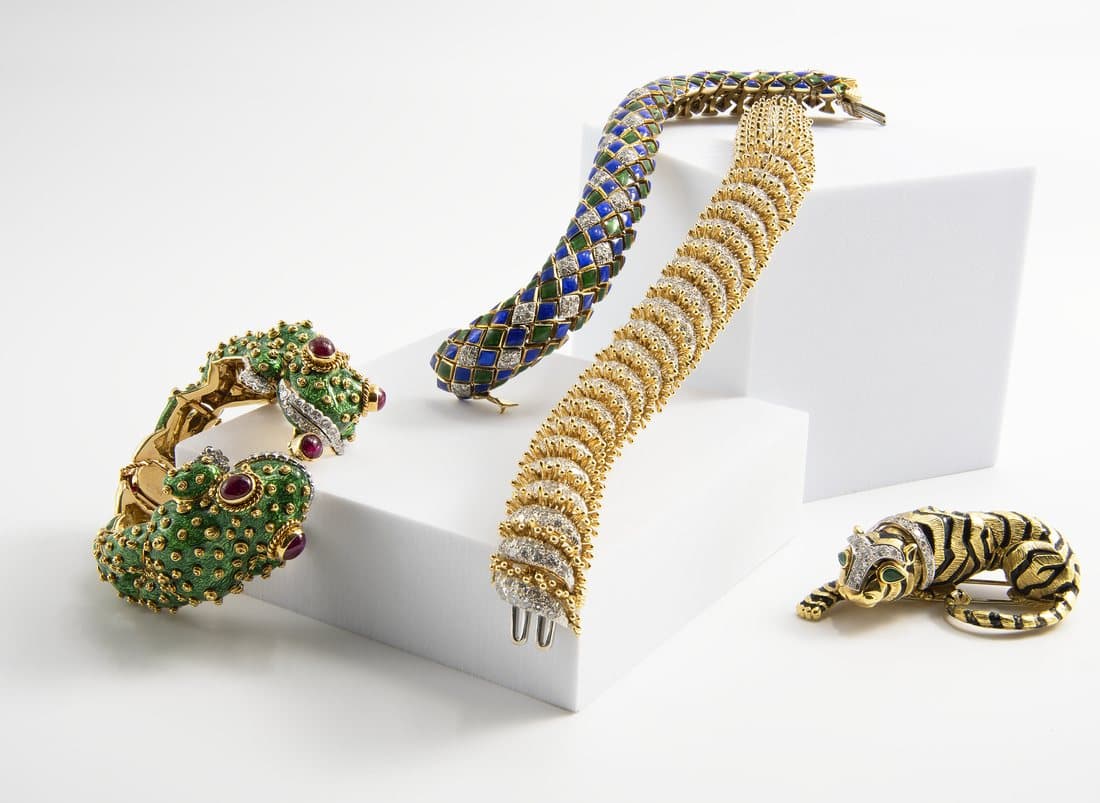 Four ornate designer pieces on white geometric pedestals, including enamel animal jewelry and gold bracelets