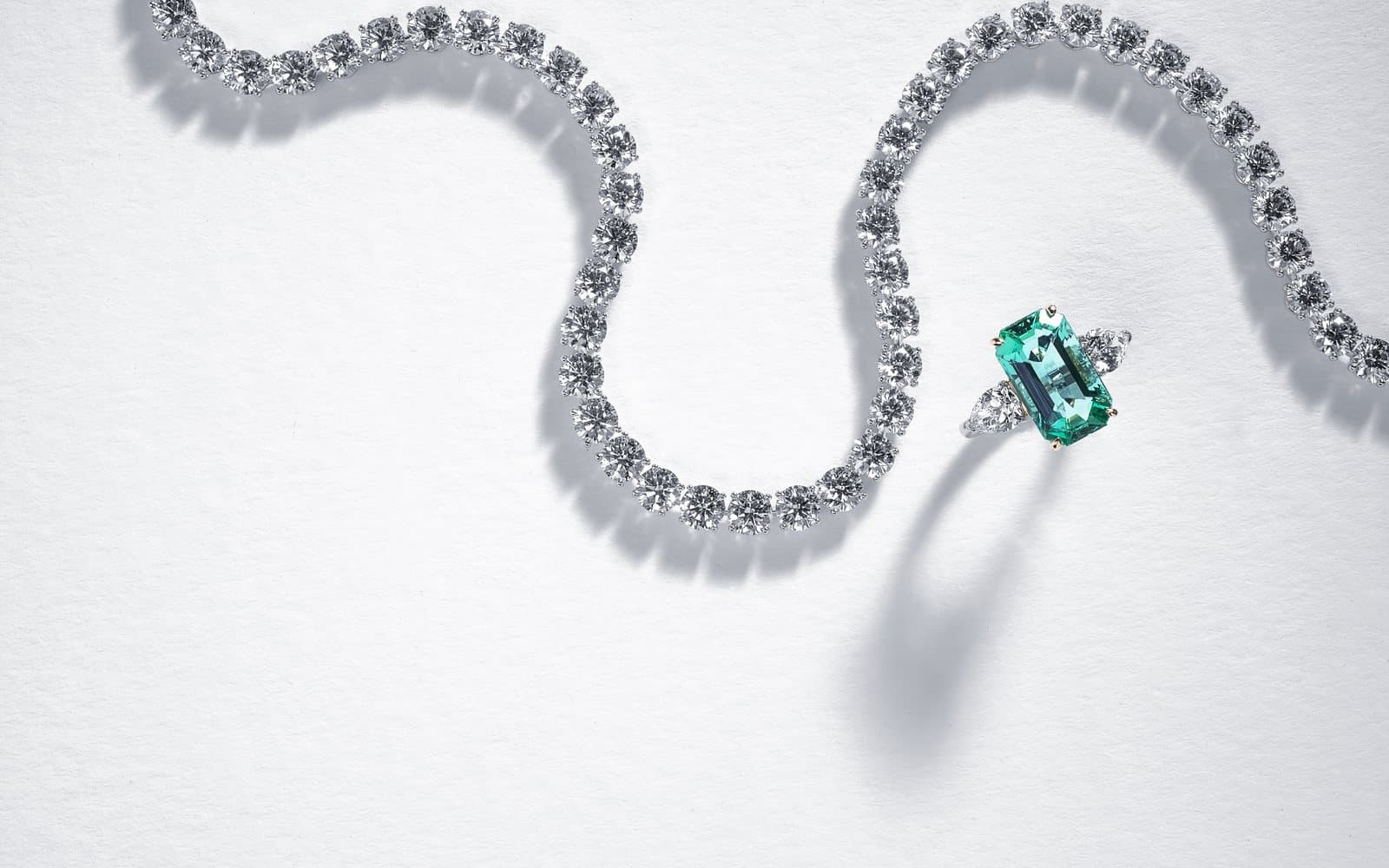 A Diamond Banc diamond riviera necklace beside an emerald-cut green gemstone and diamond ring